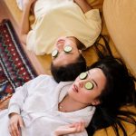 Two women enjoying a relaxing spa day with cucumber masks in a cozy home setting.